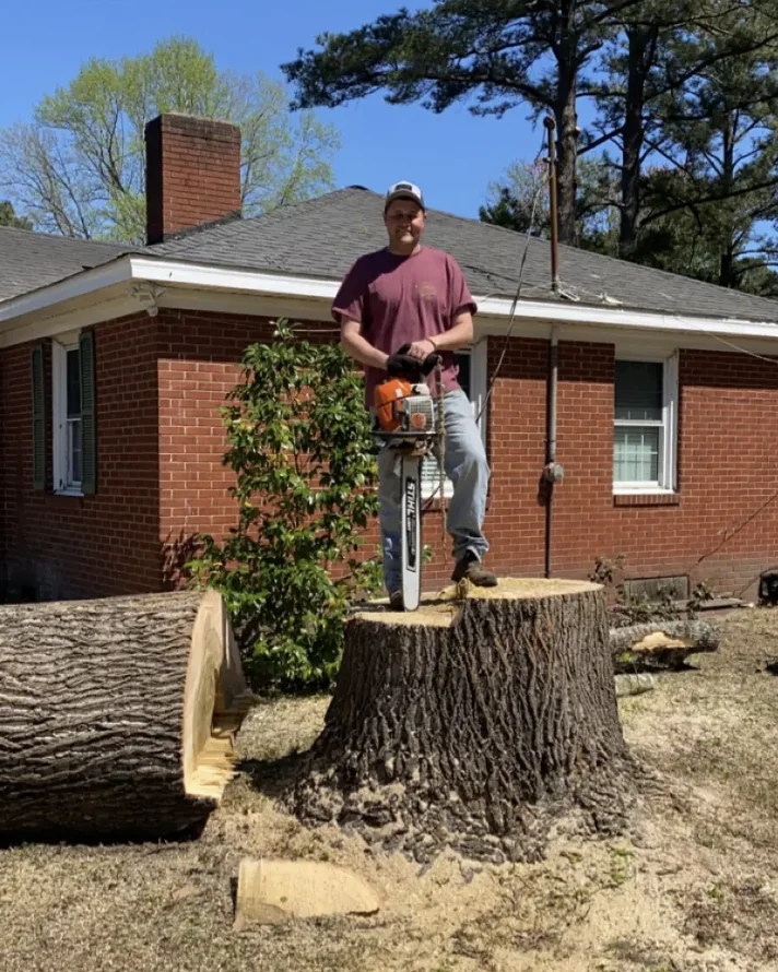 Matthew Bryan, owner of Cherry's Tree Service, holding a chainsaw on a freshly cut stump in front of a brick home in Greenville, NC